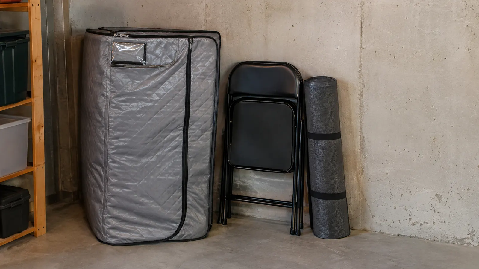 A portable steam sauna tent folded and stored against a basement wall with the chair and mat beside it, showing how little space it takes