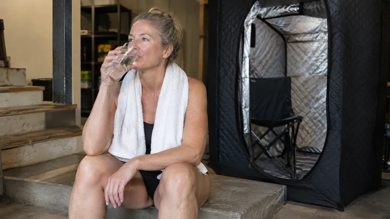 A woman in her fifties sitting on a basement step with a towel around her shoulders and a glass of water in her hand, a portable steam sauna tent open behind her